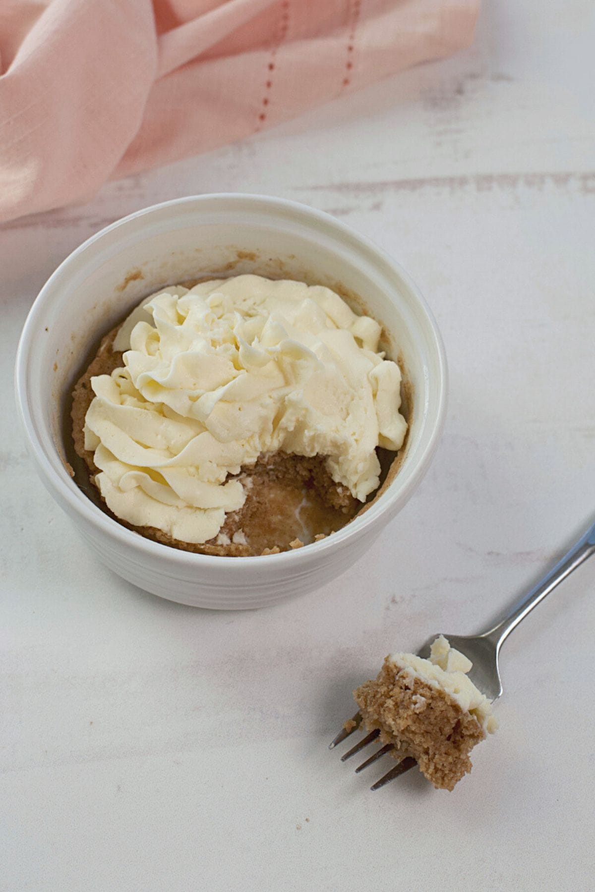 A ramekin with a partially eaten cake in mug made from cake mix, topped with whipped cream, is placed on a white surface. A fork with a piece of the dessert rests next to it. A pink fabric is visible in the background.