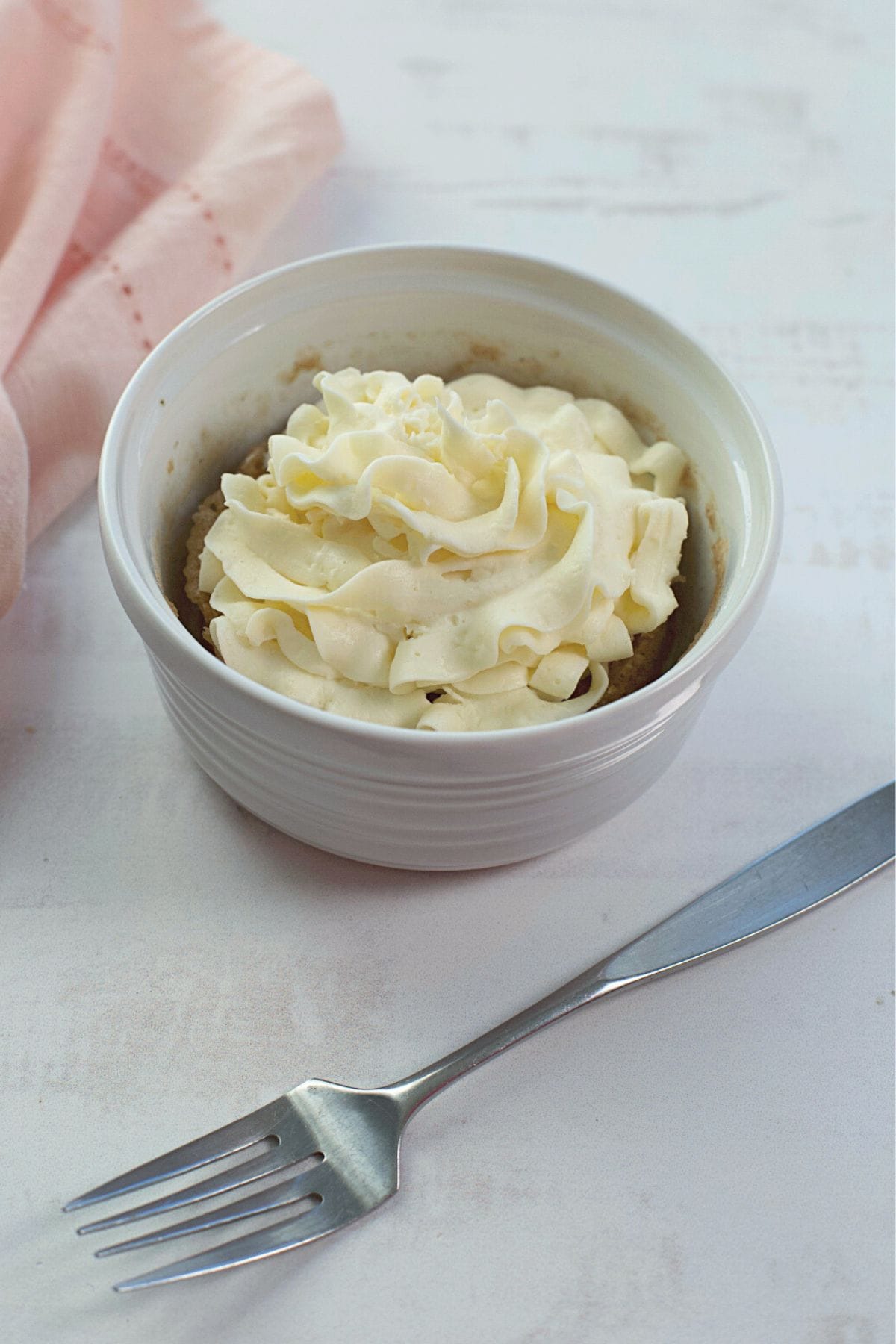 A small ceramic bowl filled with a dessert made from cake mix, topped with whipped cream, sits on a white surface next to a fork and a pink cloth.