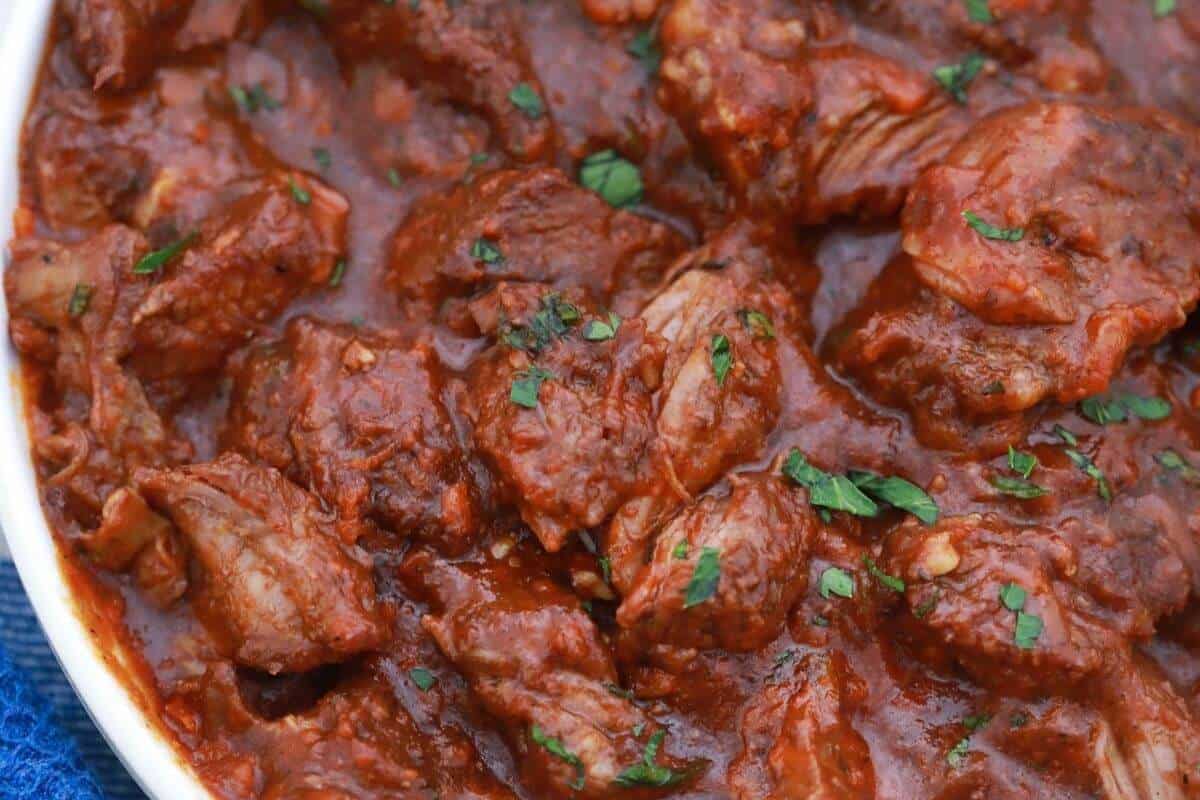 Close-up of braised short ribs and beef chunks in a rich red tomato-based sauce, garnished with chopped herbs.
