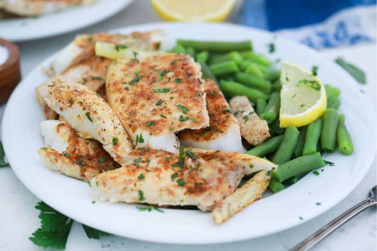 Grilled butter fish fillets garnished with herbs, served with green beans and a lemon wedge on a white plate.