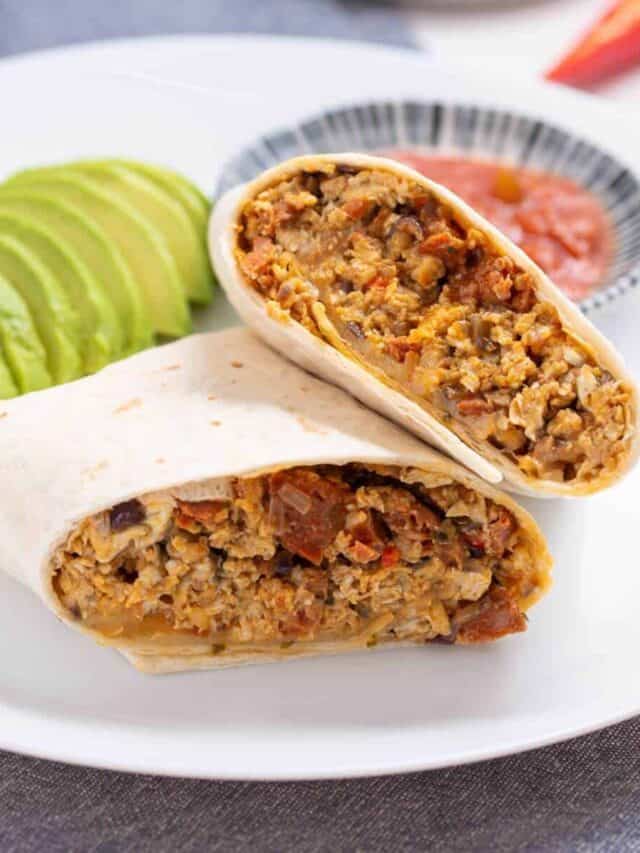 A breakfast burrito cut in half, filled with scrambled eggs and vegetables, is on a white plate with slices of avocado and a bowl of salsa in the background.