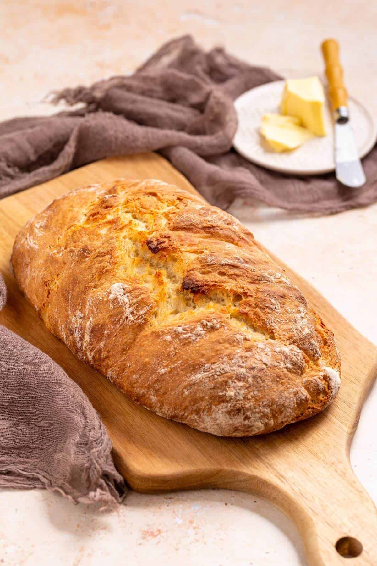 No knead Italian artisan bread loaf on cutting board with butter in back.