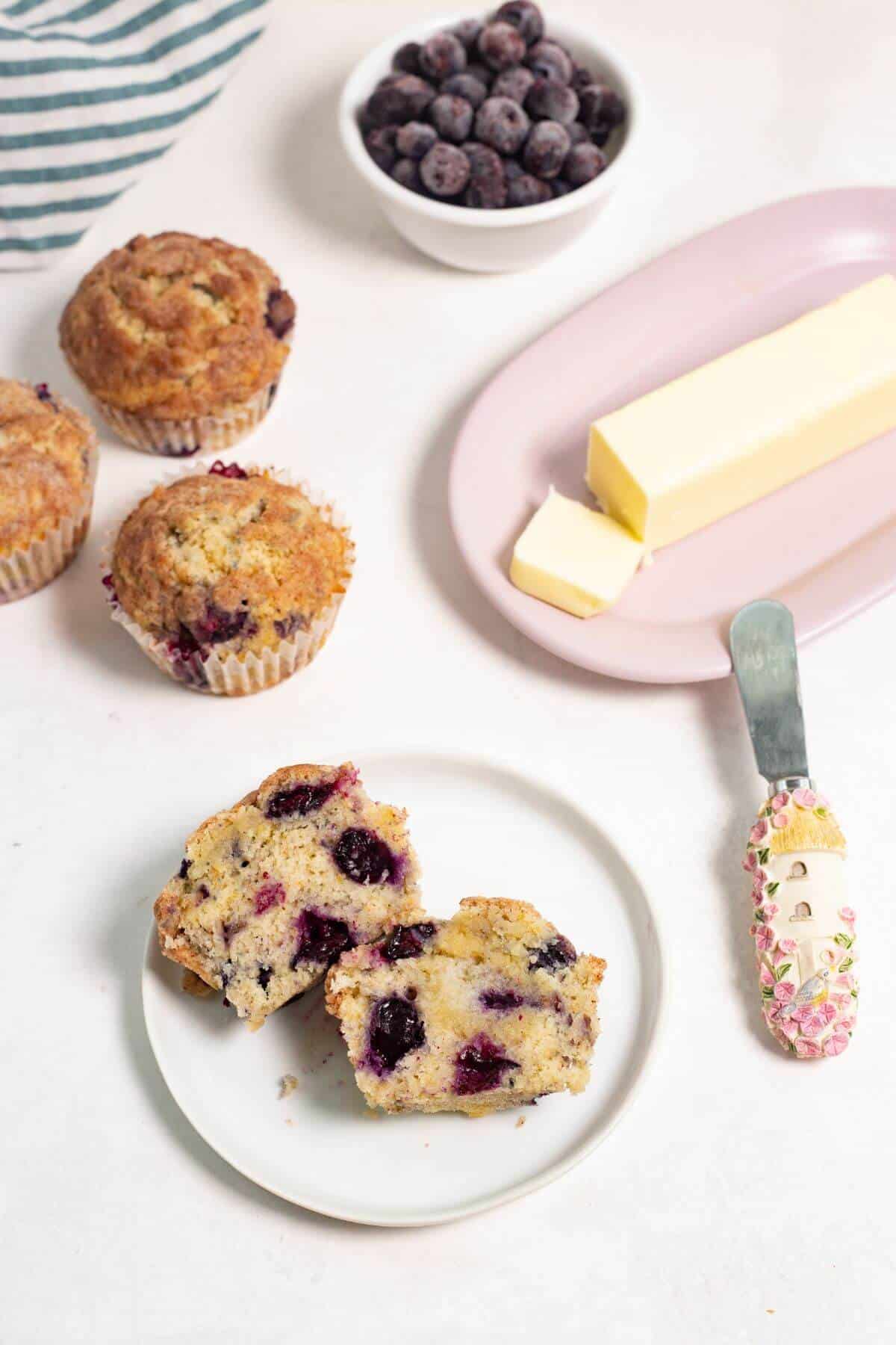 Sliced blueberry muffin on plate with butter, blueberries, and other muffins.