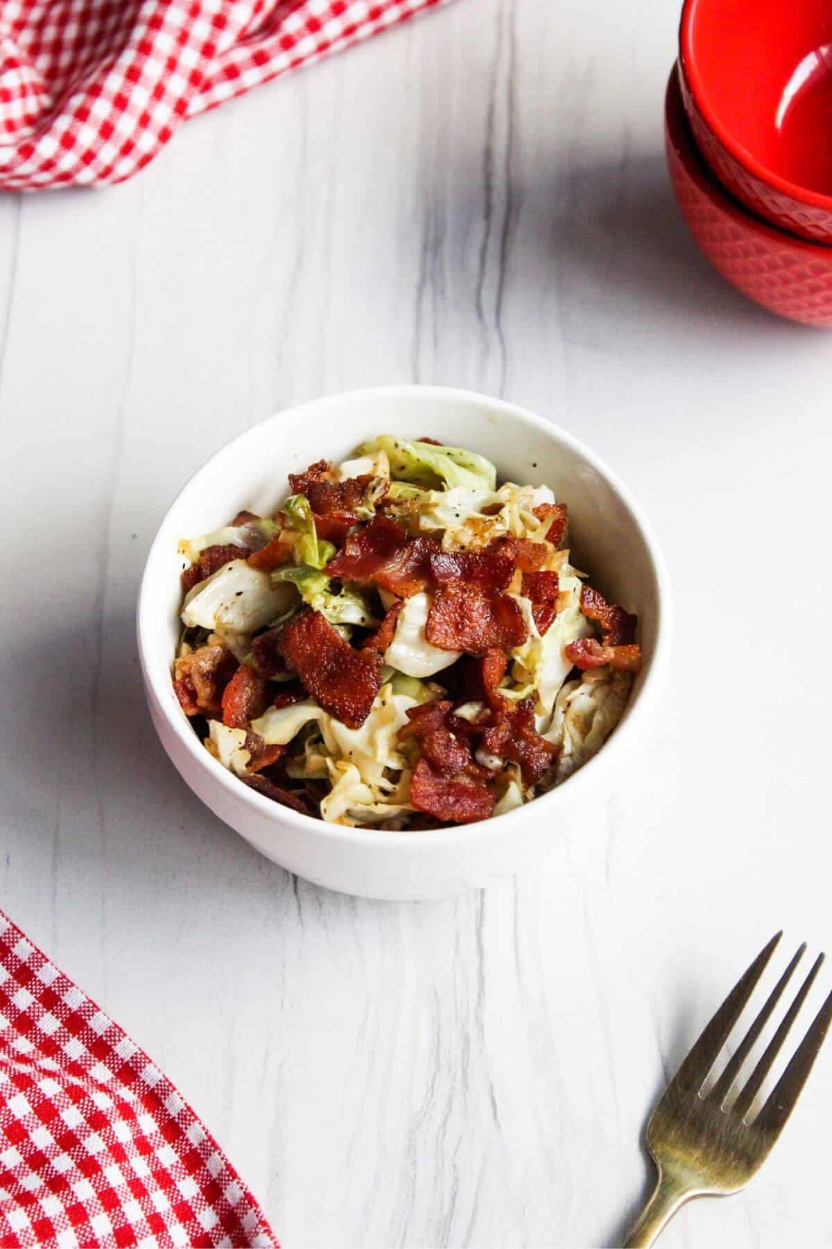 Southern fried cabbage in white bowl with red bowls and fork on sides.