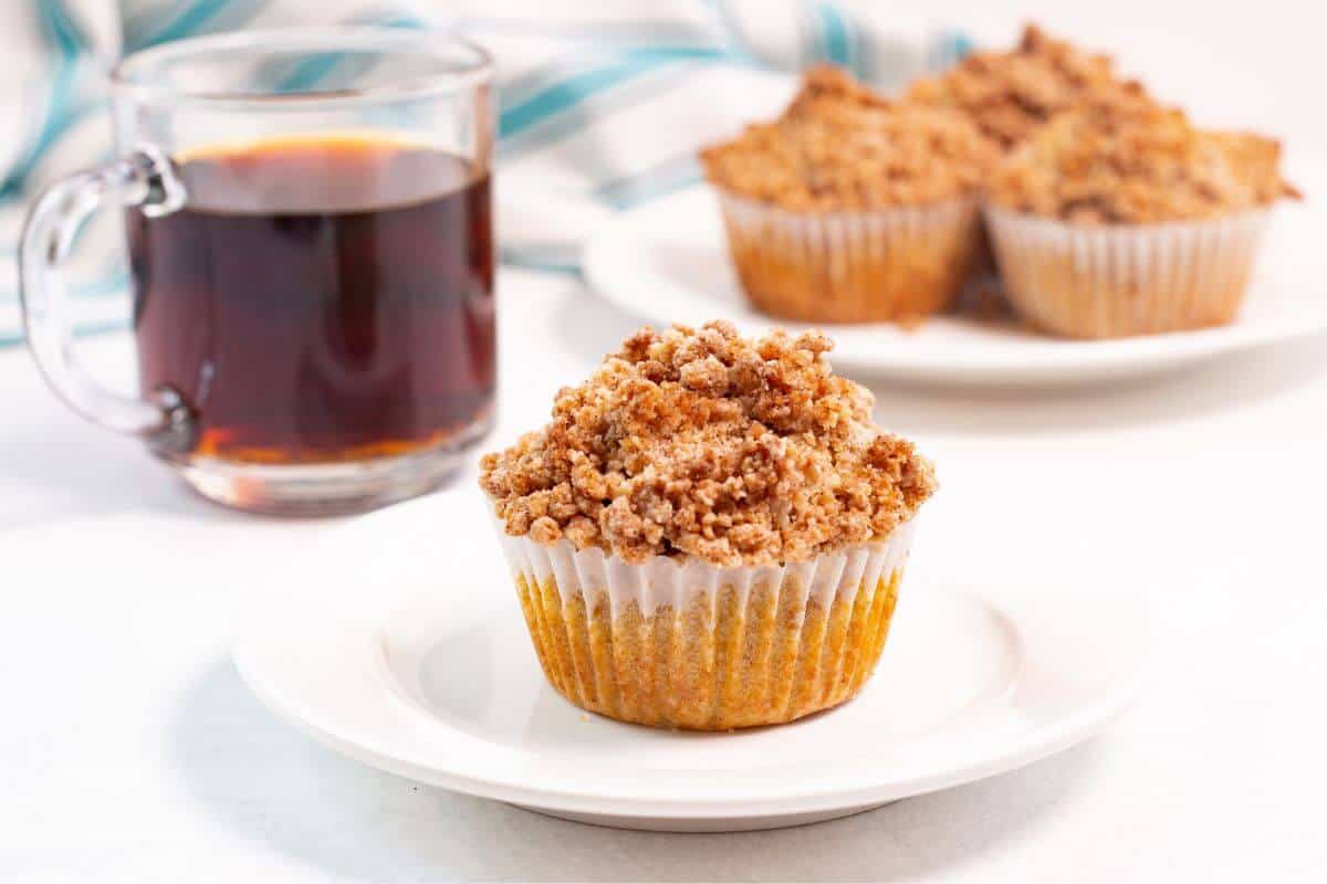Coffee cake muffin on plate with coffee and more muffins in back.