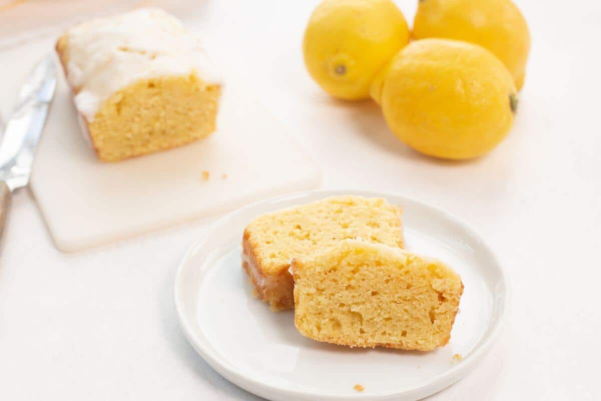 Lemon loaf slices on small plate with lemons and loaf in back.