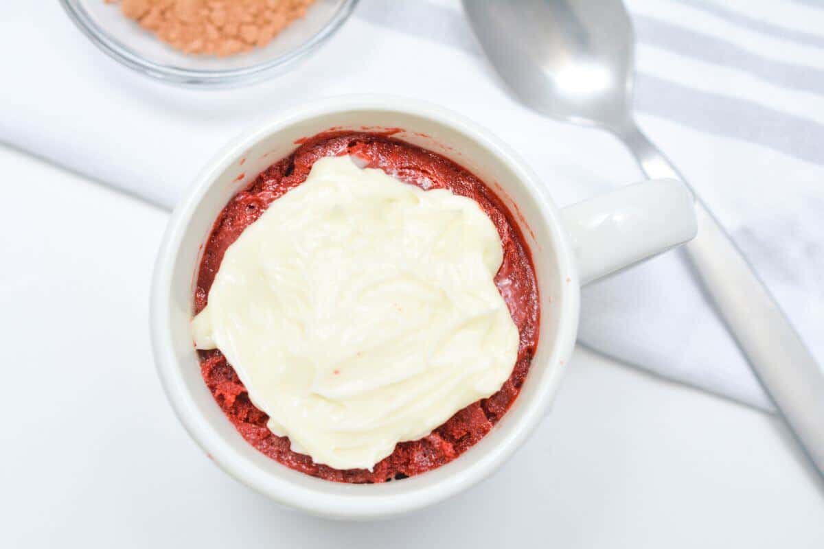 Frosted red velvet mug cake with spoon and cocoa.