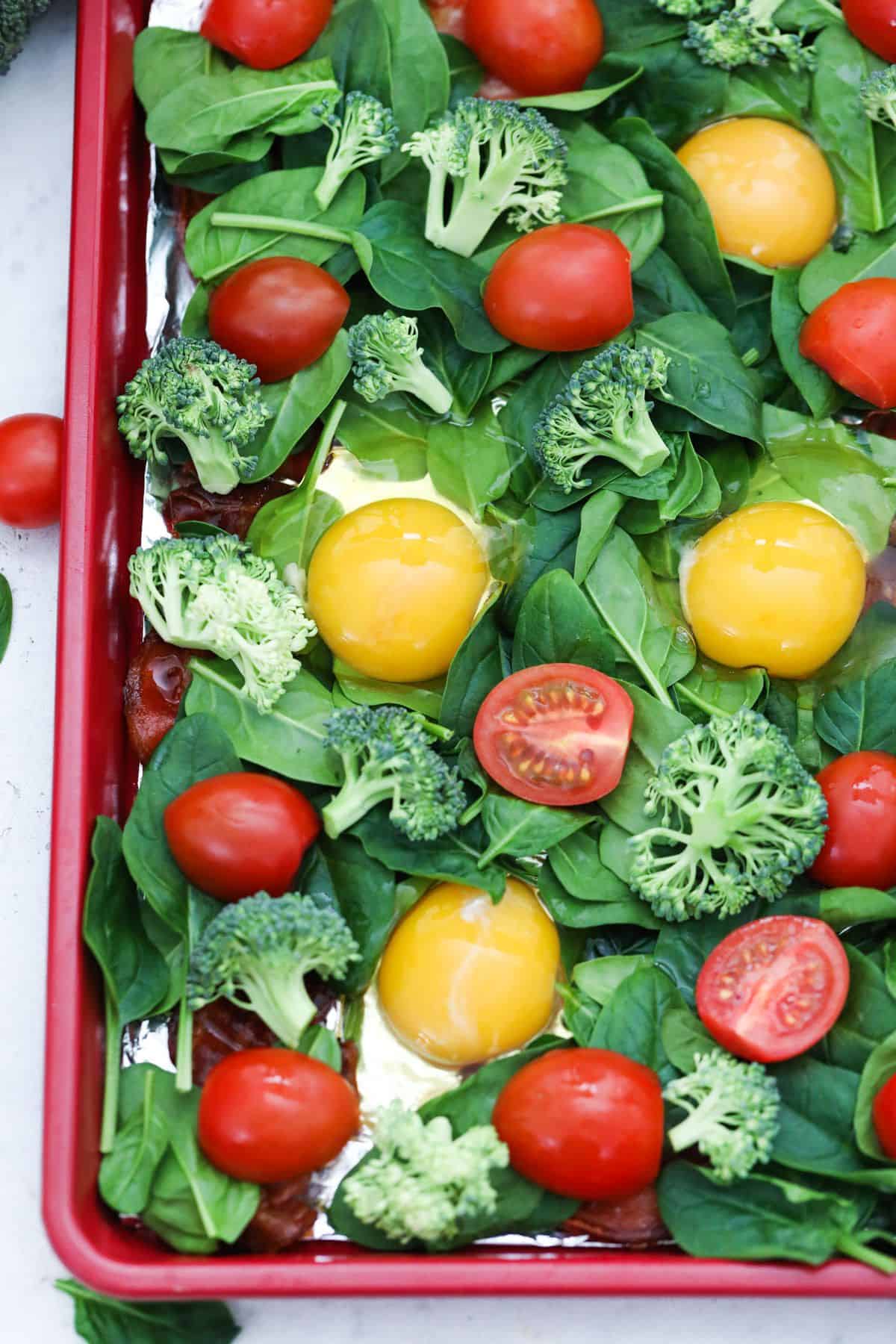 Spinach, broccoli, eggs, and cherry tomatoes over bacon in sheet pan.