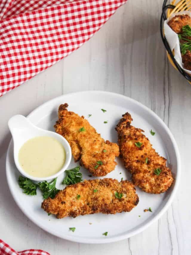 Three breaded chicken tenders on a white plate with a side of dipping sauce, garnished with parsley, on a wooden table with a red checkered napkin.