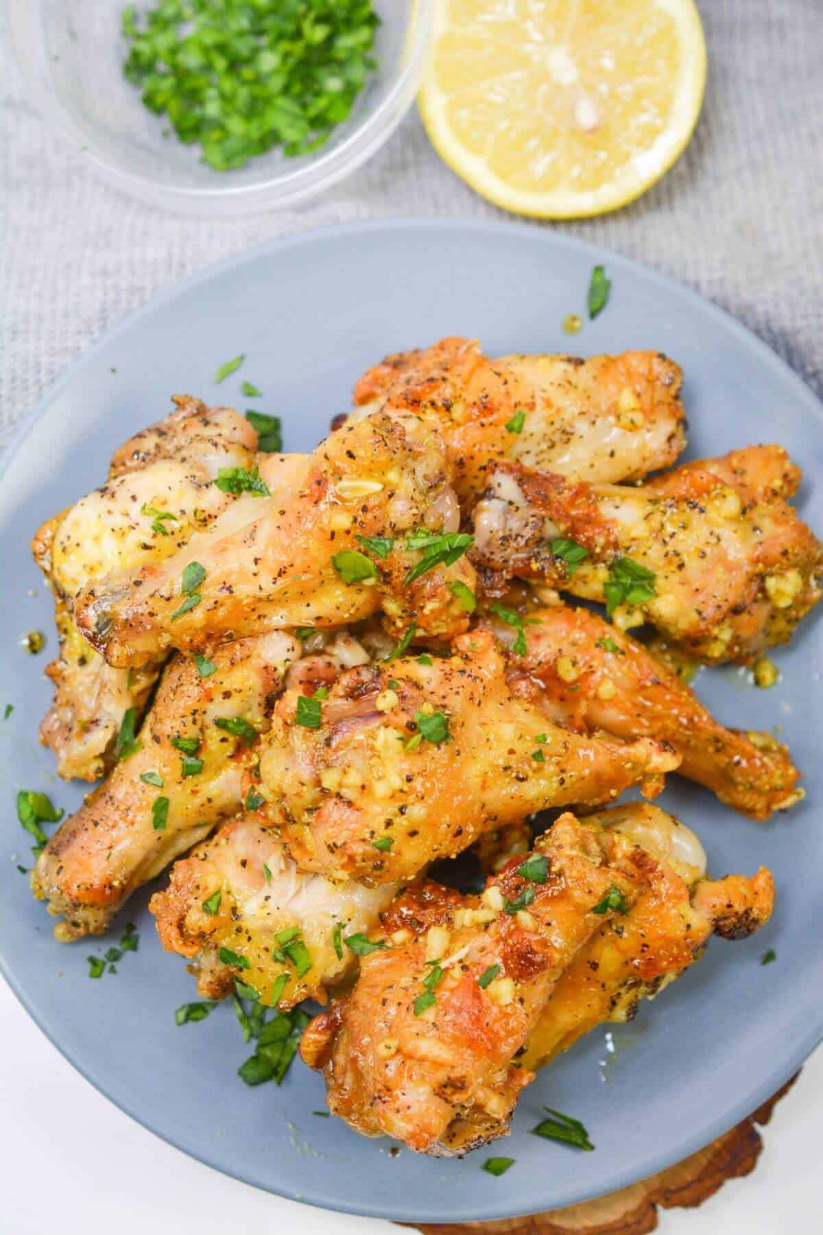 A plate of seasoned lemon pepper chicken wings garnished with chopped herbs, next to a lemon half and a small bowl of green herbs.
