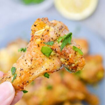 A hand holding a seasoned, cooked lemon pepper chicken wing garnished with chopped parsley, with more lemon pepper chicken wings blurred in the background.