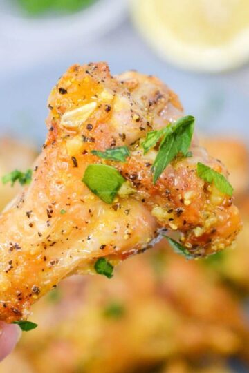 A hand holding a seasoned, cooked lemon pepper chicken wing garnished with chopped parsley, with more lemon pepper chicken wings blurred in the background.