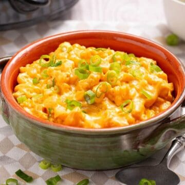 Bowl of creamy crock pot mac and cheese topped with chopped green onions, placed on a table with a spoon nearby.
