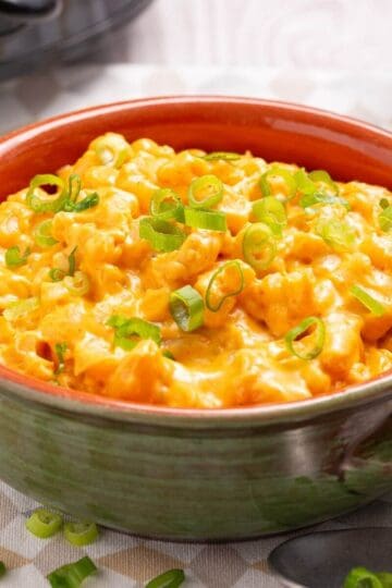 Bowl of creamy crock pot mac and cheese topped with chopped green onions, placed on a table with a spoon nearby.