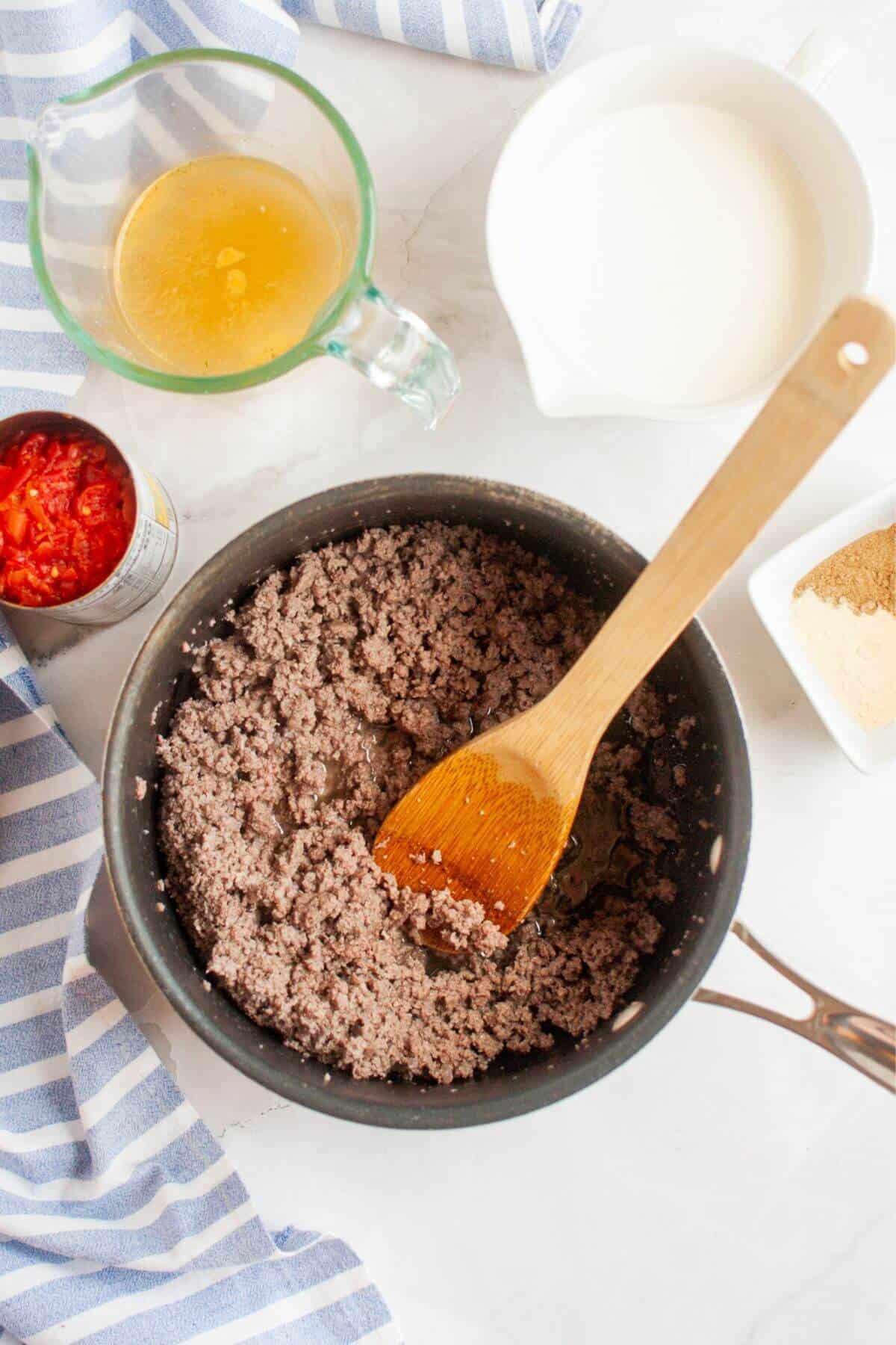 A pan with cooked ground beef queso dip and a wooden spoon, surrounded by a measuring cup with broth, a pitcher of milk, an open can of tomatoes, and a dish of seasoning. .