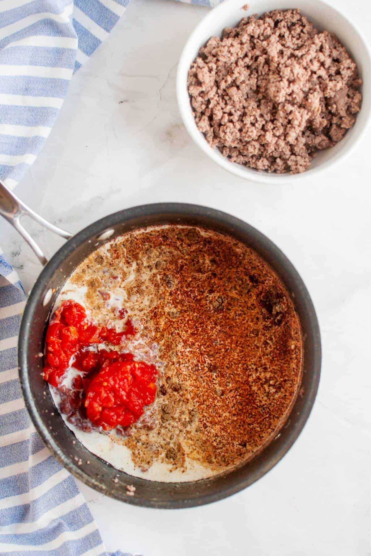 A pot with milk, beef queso dip, and tomato sauce rests on a marble surface next to a bowl of cooked ground meat.