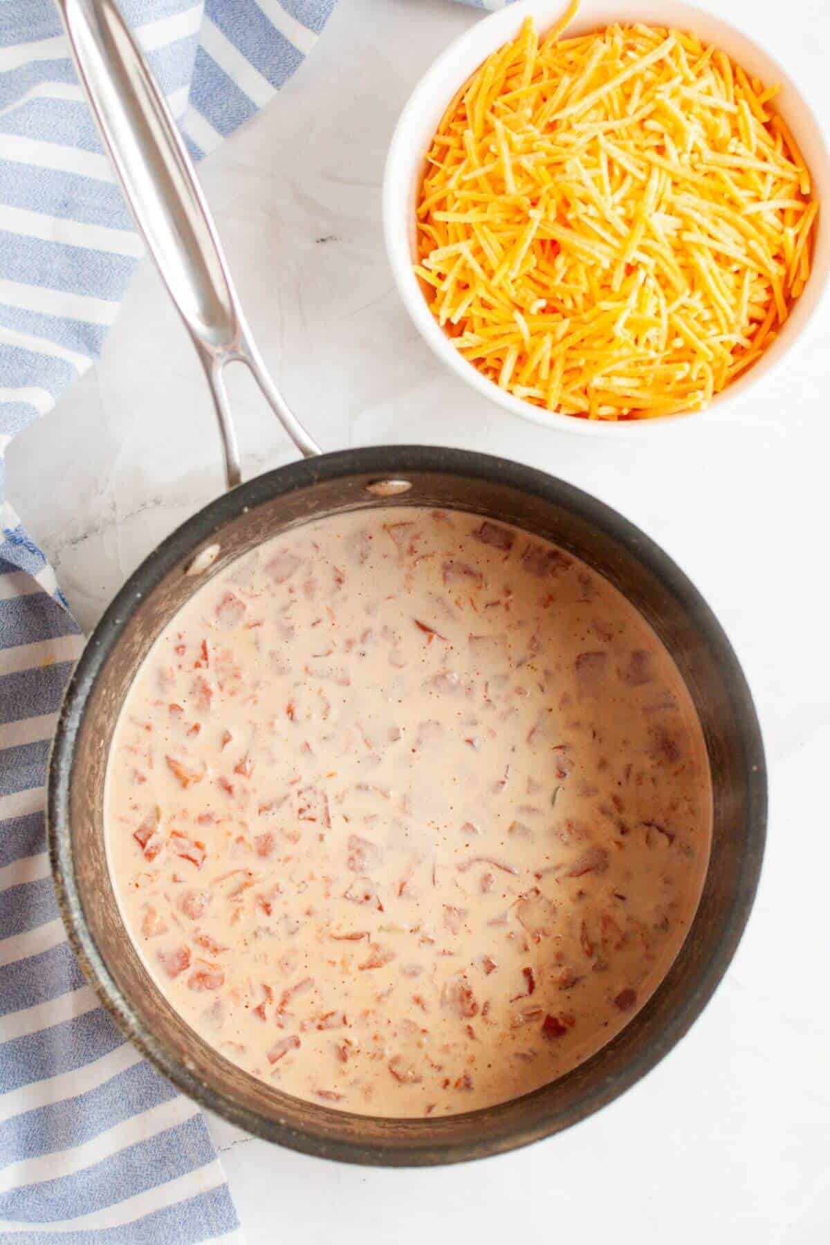 A pot of creamy tomato soup sits enticingly next to a bowl of shredded cheese on a striped cloth, reminiscent of the rich flavors found in a classic beef queso dip.