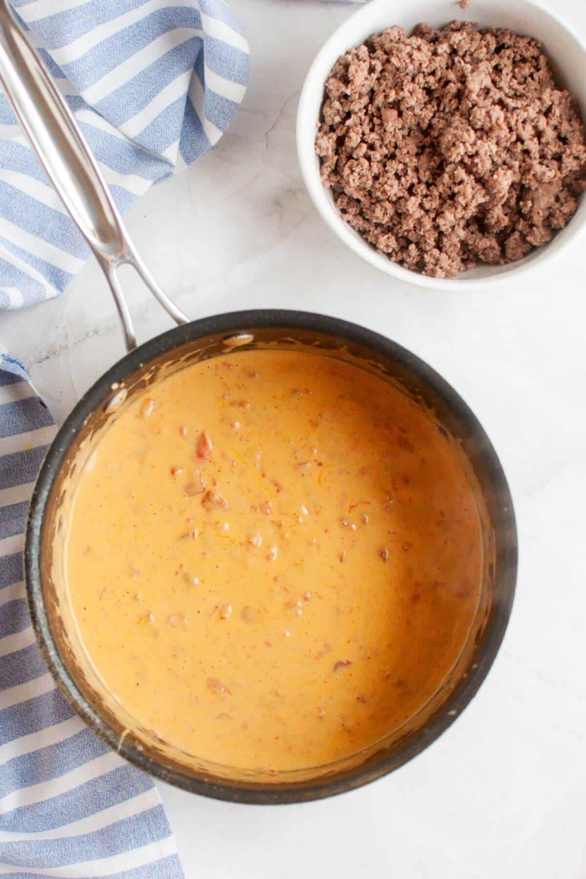 A saucepan filled with creamy cheese sauce and a white bowl containing cooked ground beef create the perfect ingredients for a delicious beef queso dip on a marble surface, accented by a blue striped cloth nearby.