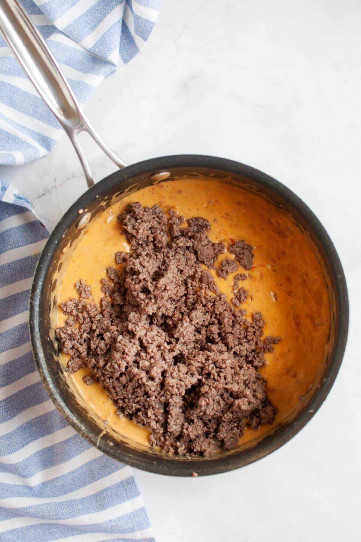 A saucepan contains a delicious beef queso dip with a mix of ground beef and melted cheese, resting beside a blue and white striped cloth.