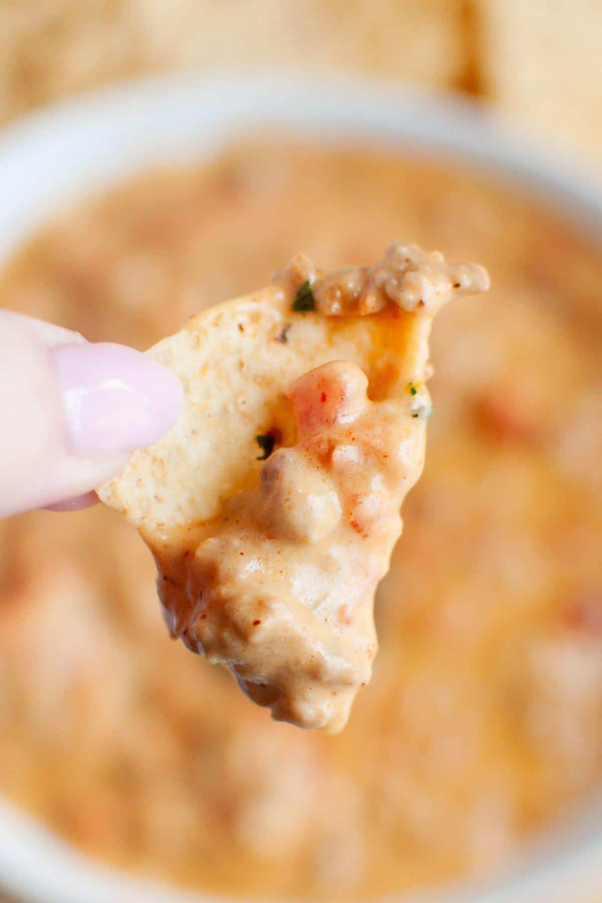 A close-up of a hand holding a tortilla chip dipped in rich beef queso, with cheesy goodness and chunks of tomato and herbs.