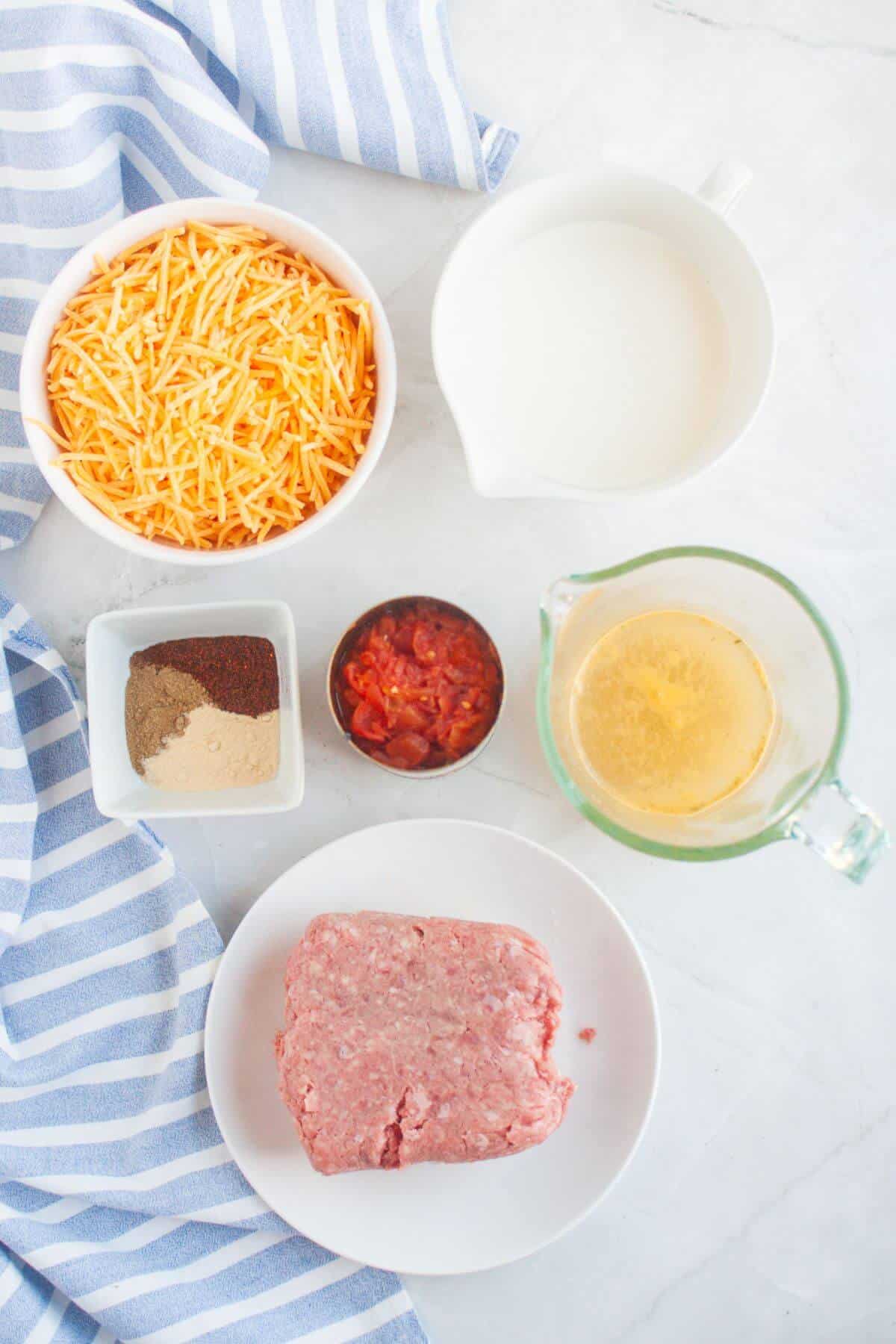 Ingredients for a delicious beef queso dip on a table: shredded cheese, milk, ground meat, spices in a small dish, diced tomatoes in a bowl, and broth in a measuring cup. A striped cloth lies underneath.