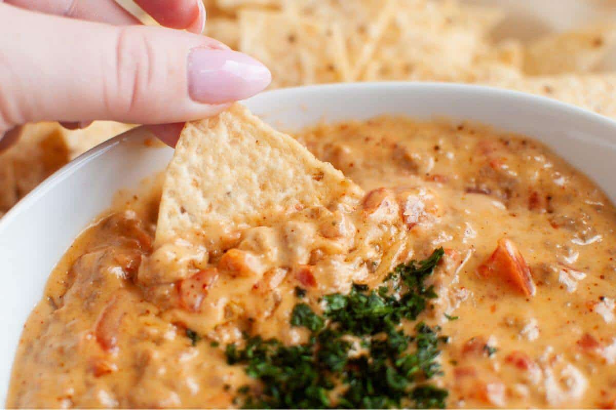 A hand dips a tortilla chip into a bowl of cheesy beef queso dip with herbs.
