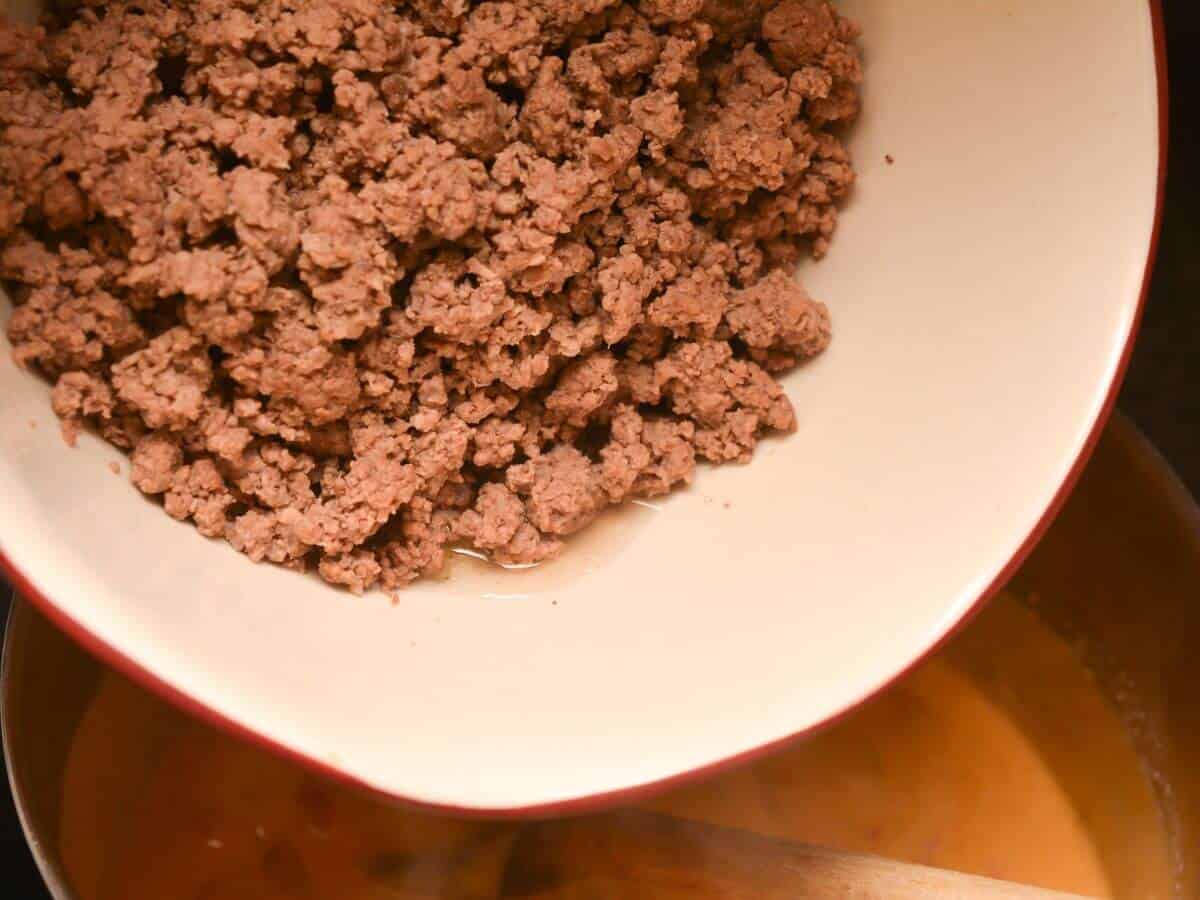 A bowl of ground beef being poured into cheese mixture in pot.