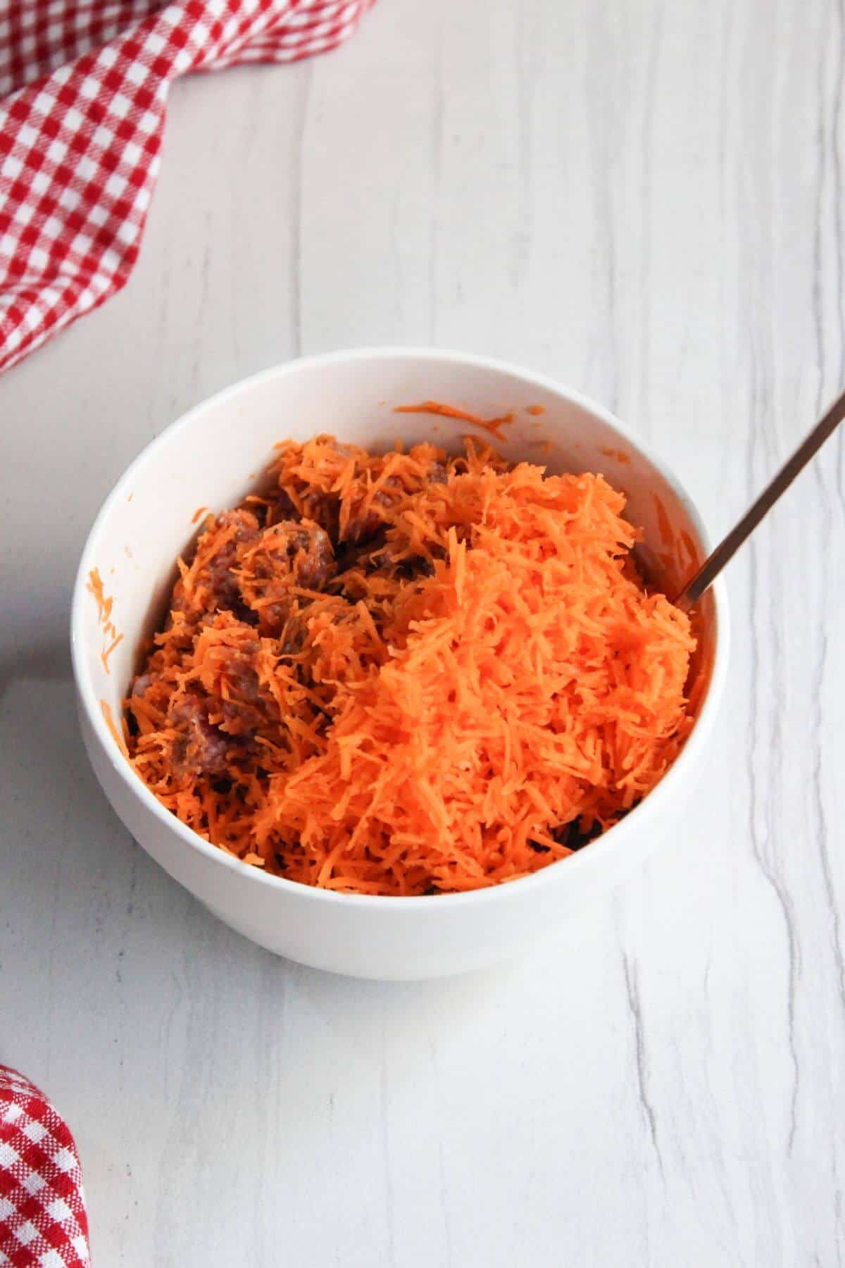 Carrots added to meat in a white bowl with a spoon.