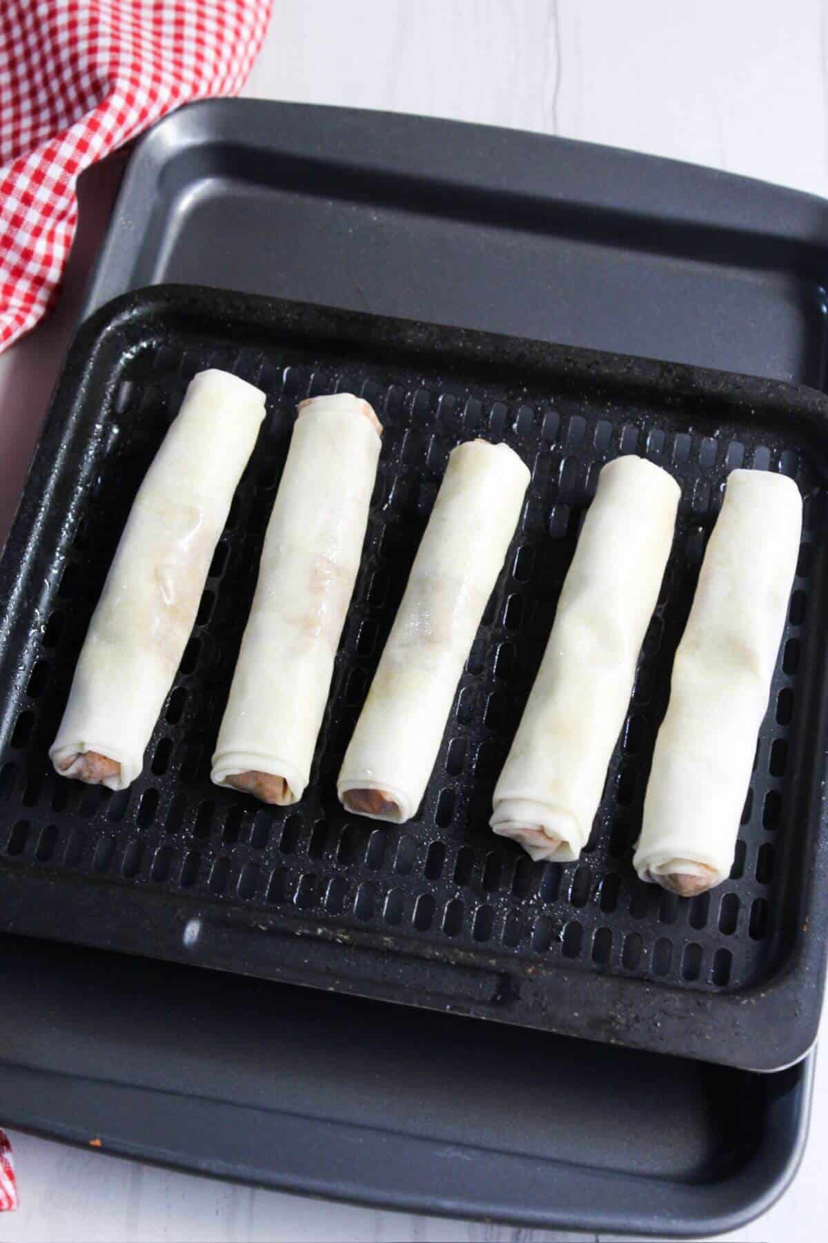 Uncooked lumpia rolls in an air fryer pan with a checkered checkered tablecloth.