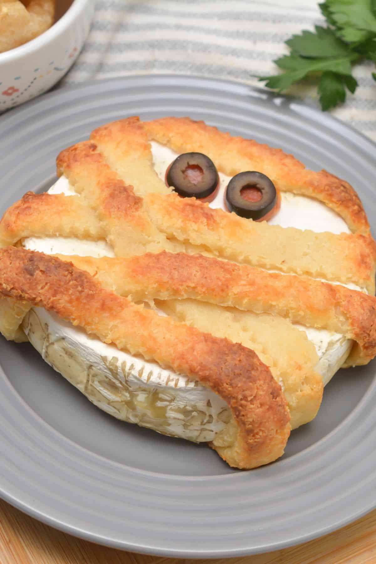 A mummy brie cheese with olive eyes on a plate.