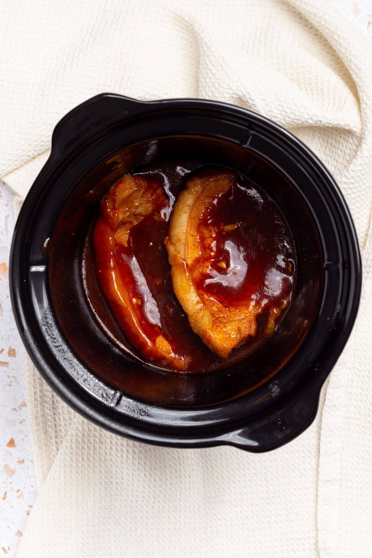 A black crock pot with pork chops and sauce.