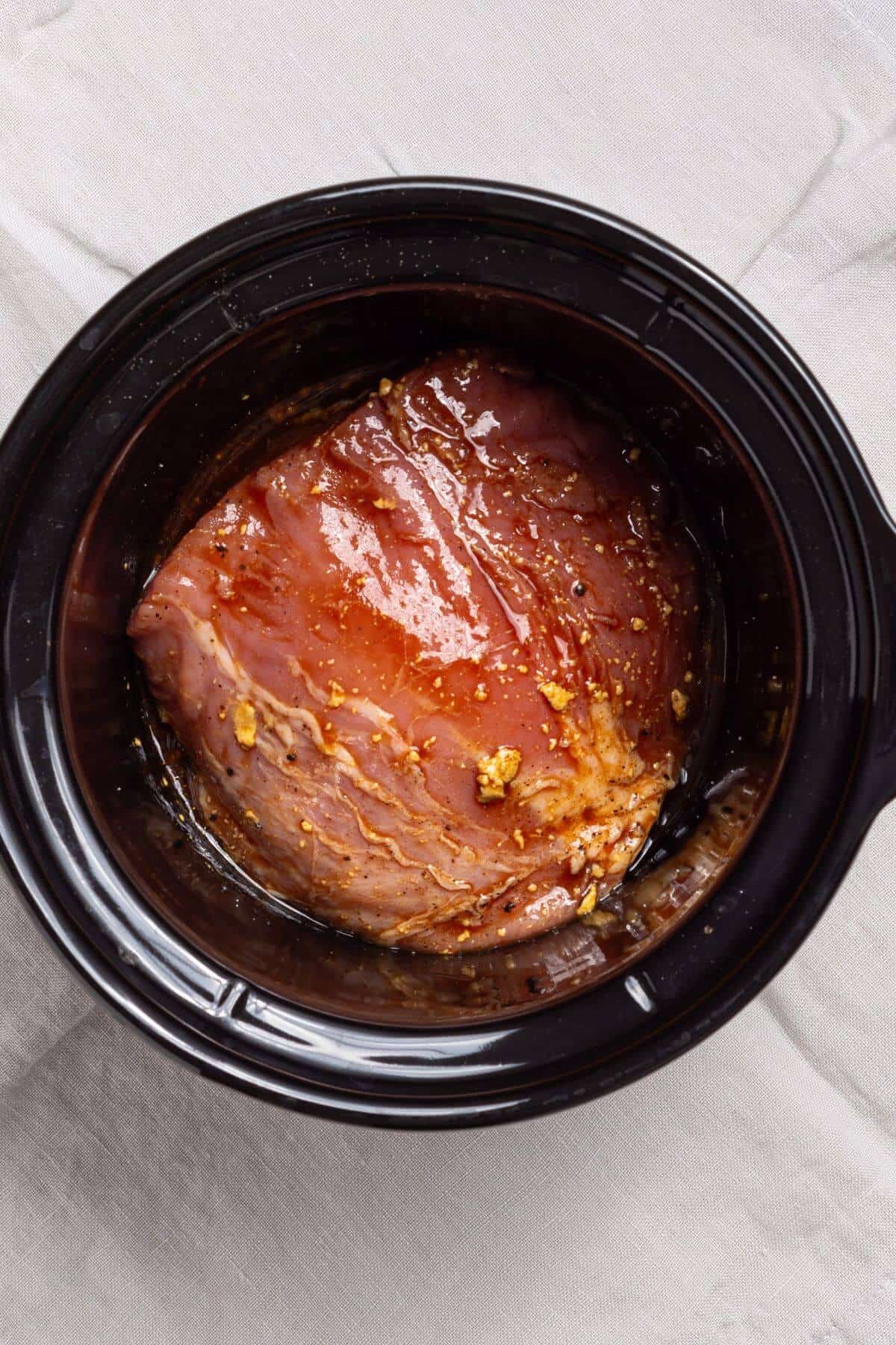 Sauce poured over pork loin in a slow cooker on a white cloth.