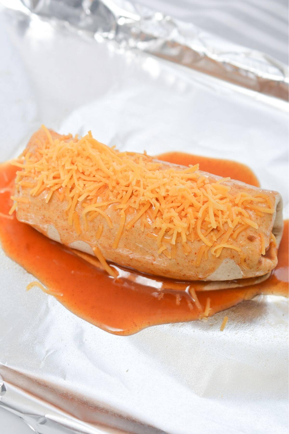 A burrito wrapped in foil with sauce and cheese.