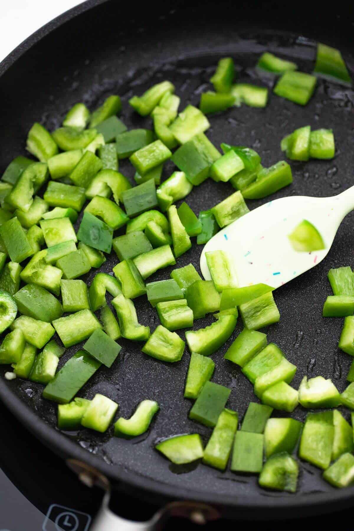 Chopped green pepper in a frying pan with a spoon.