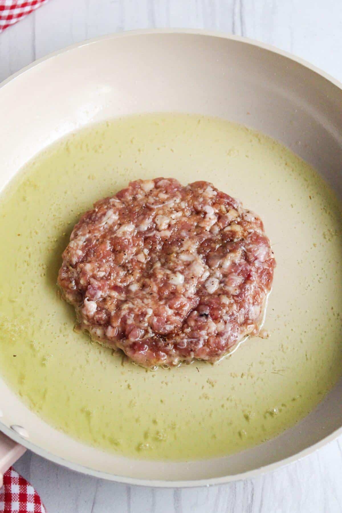A raw ground pork burger in a frying pan with oil.