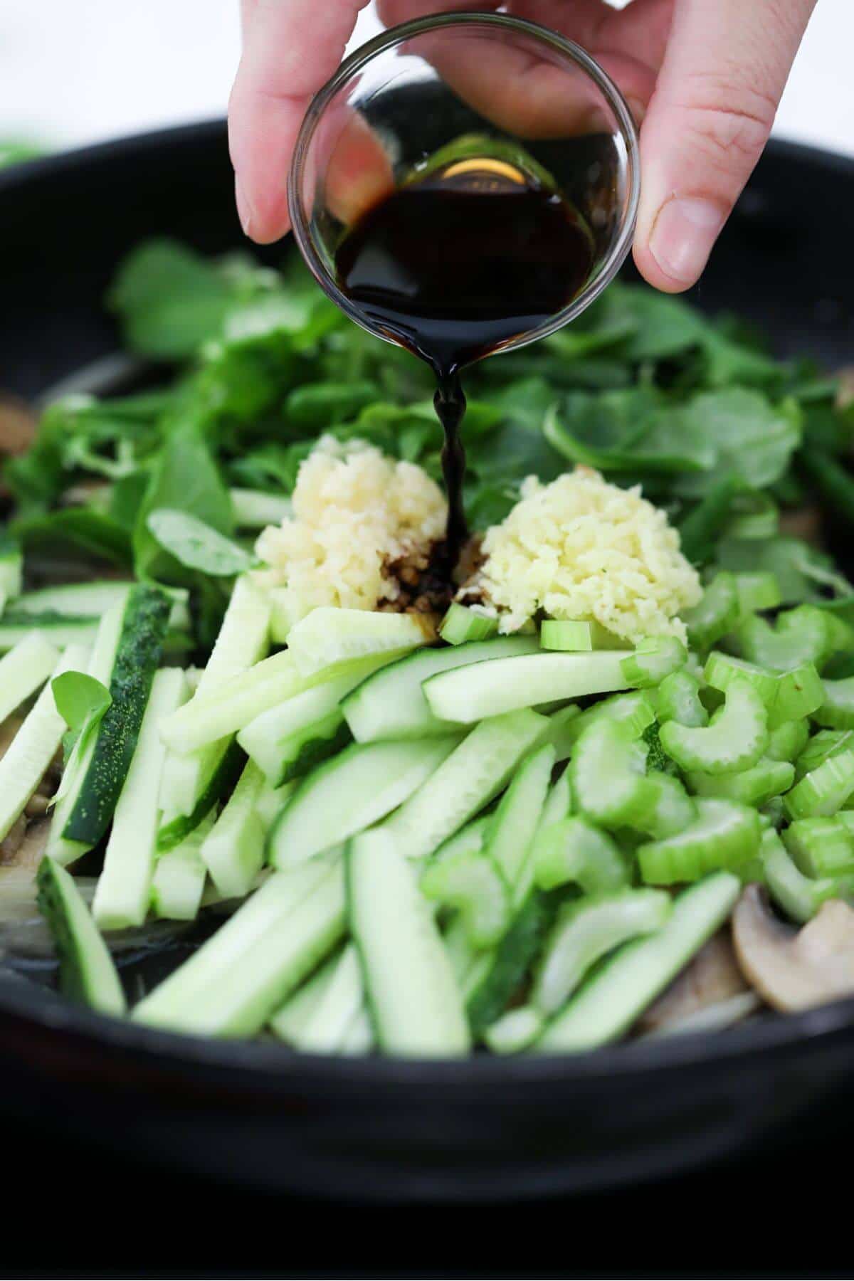 A person pouring soy sauce over vegetables in a pan.