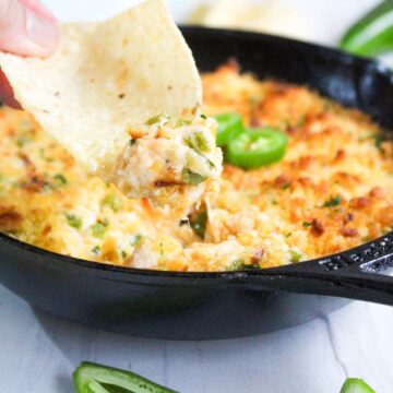 A person is dipping a tortilla chip into a skillet of cheese and jalapeno dip.