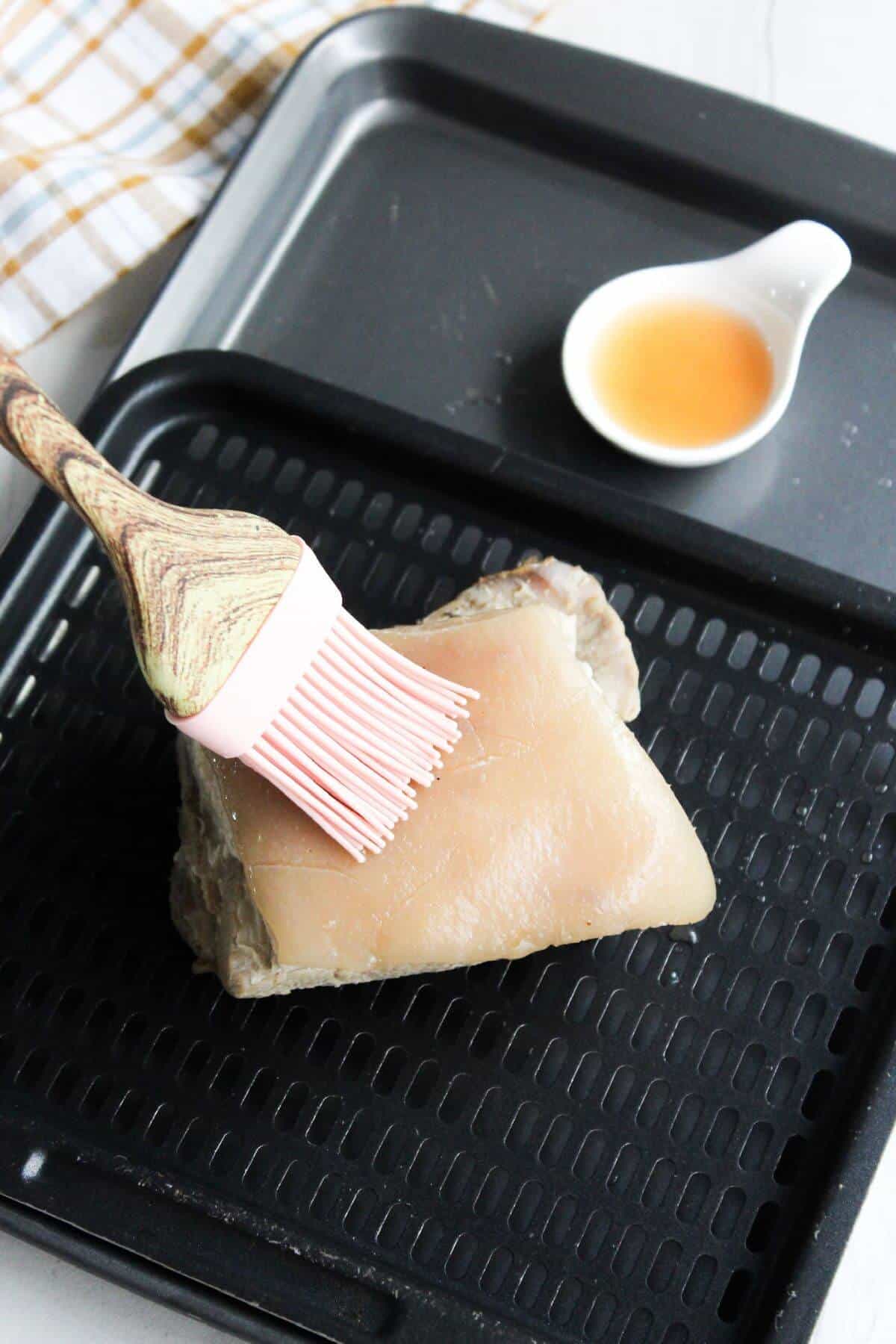 A piece of pork belly on an air fryer tray with a brush.