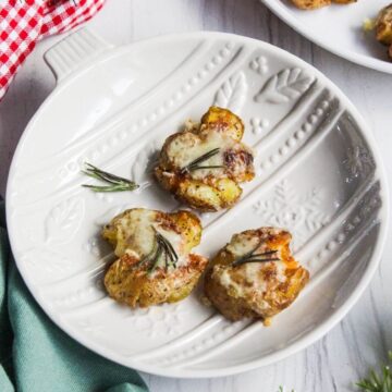 Three golden smashed potatoes topped with melted cheese and fragrant rosemary are arranged on a white decorative plate on a light surface.