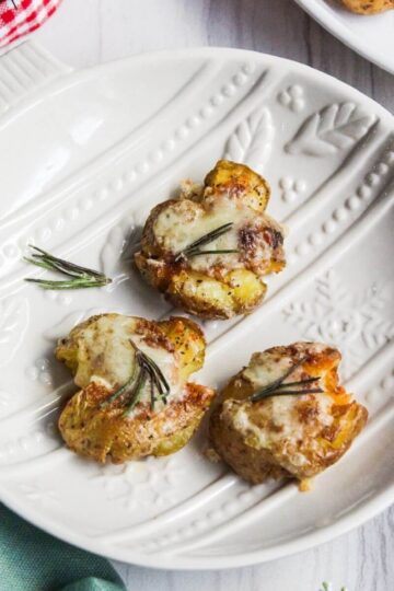 Three golden smashed potatoes topped with melted cheese and fragrant rosemary are arranged on a white decorative plate on a light surface.