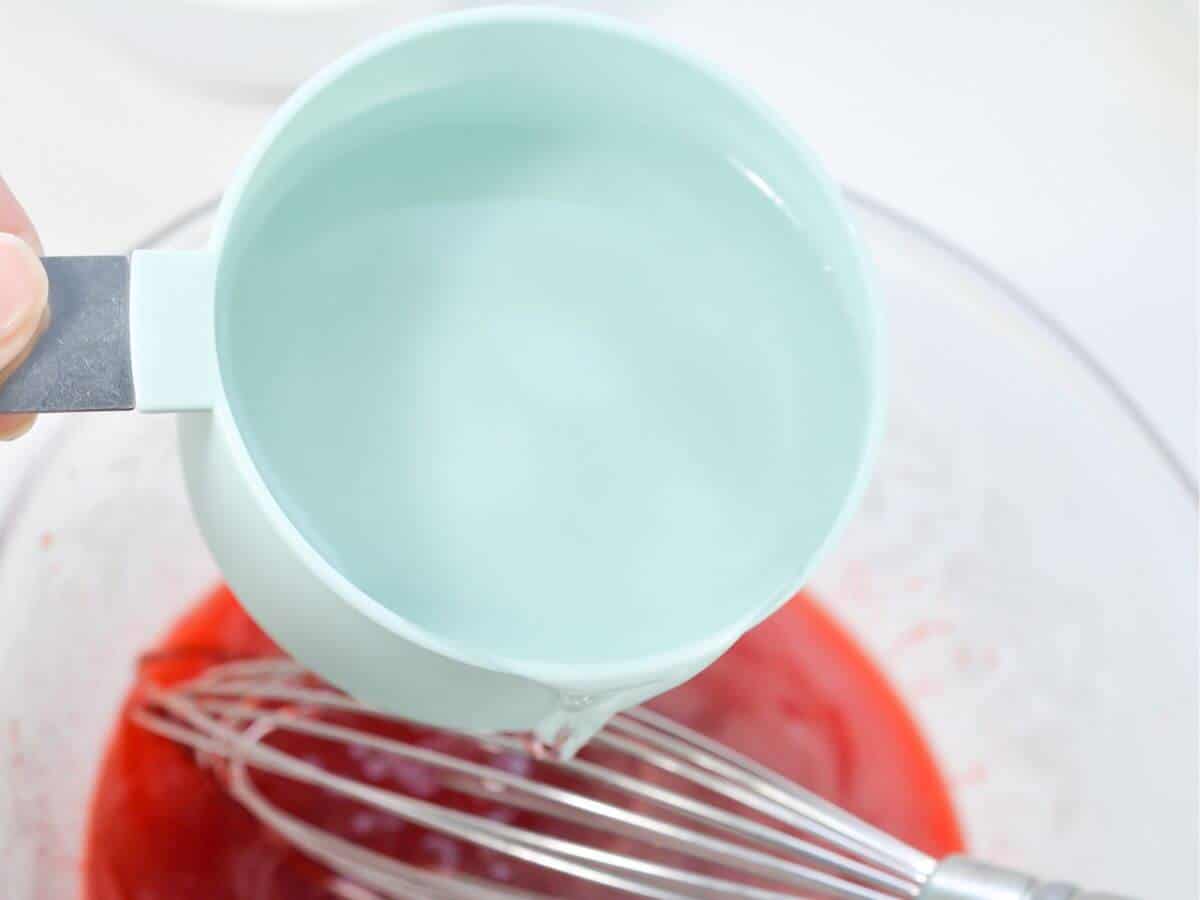 A person using a whisk to mix water into a bowl of red Jell-o mix.