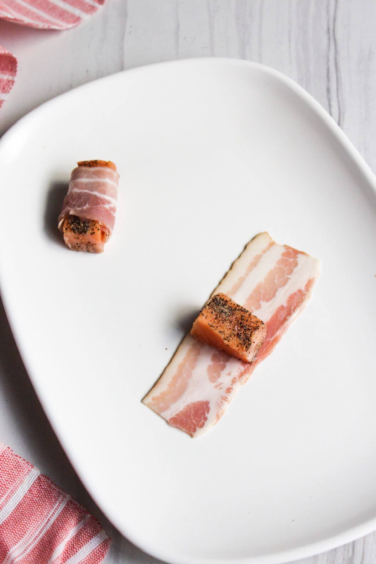 Wrapping salmon cubes in bacon on a white plate.
