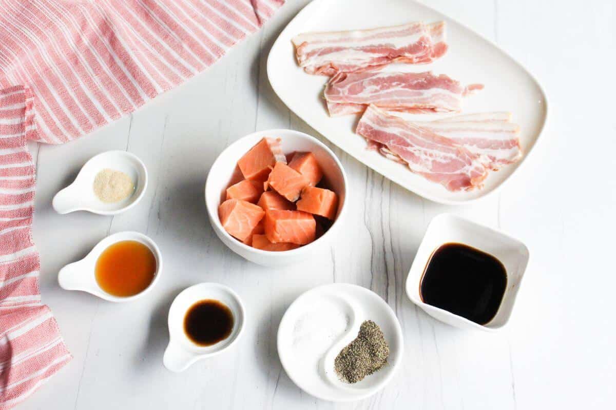 A bowl of salmon cubes, a plate of bacon slilces, sauces, and spices on a white table.
