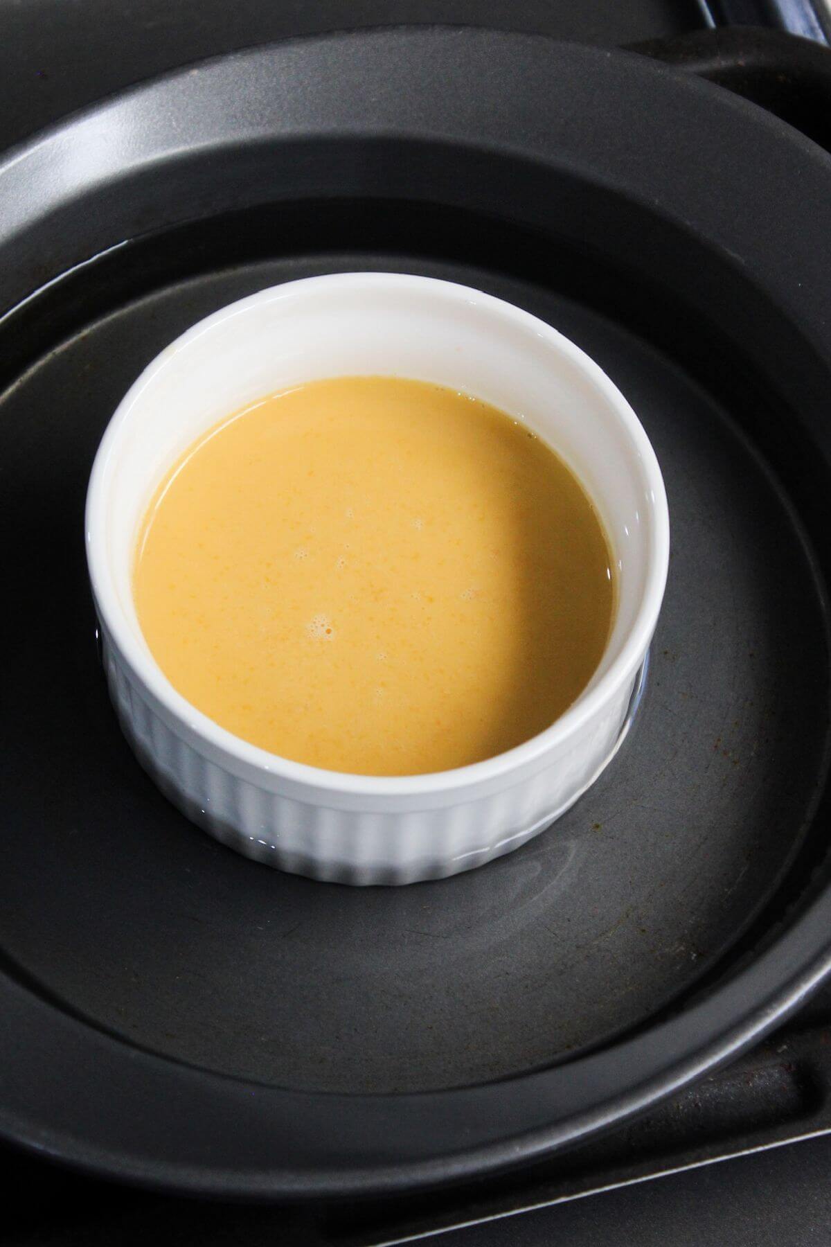A ramekin of uncooked leche flan sitting on baking pan with water bath.