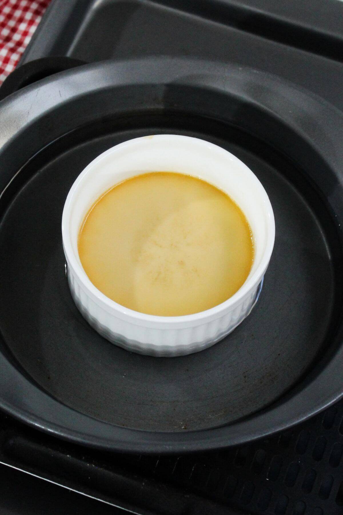 A ramekin of baked leche flan sitting on baking pan with water bath.
