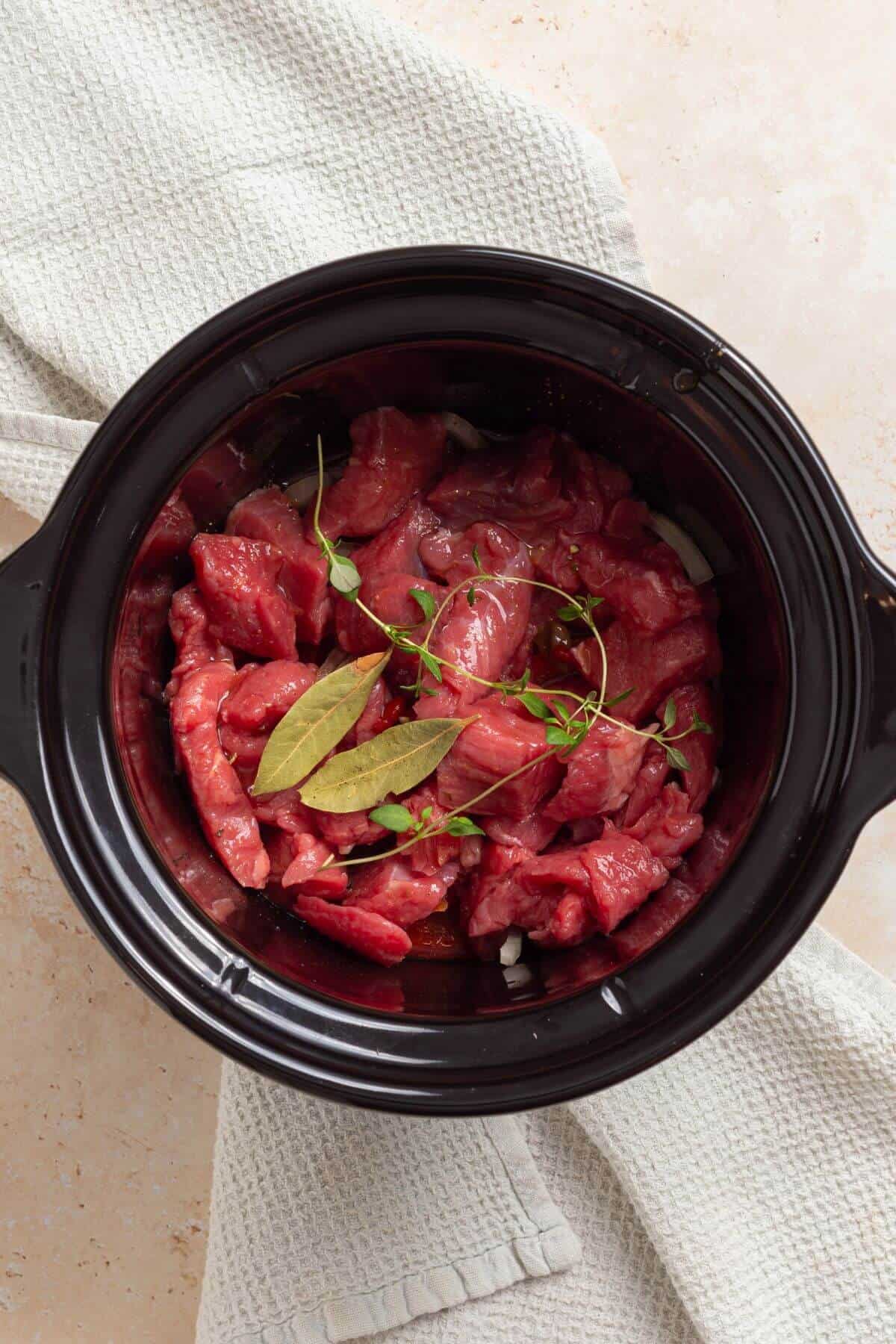 A crock pot filled with cut steak and herbs.