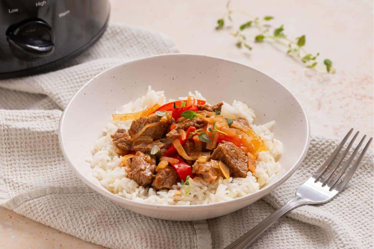 A bowl of rice with meat and peppers in front of a slow cooker.
