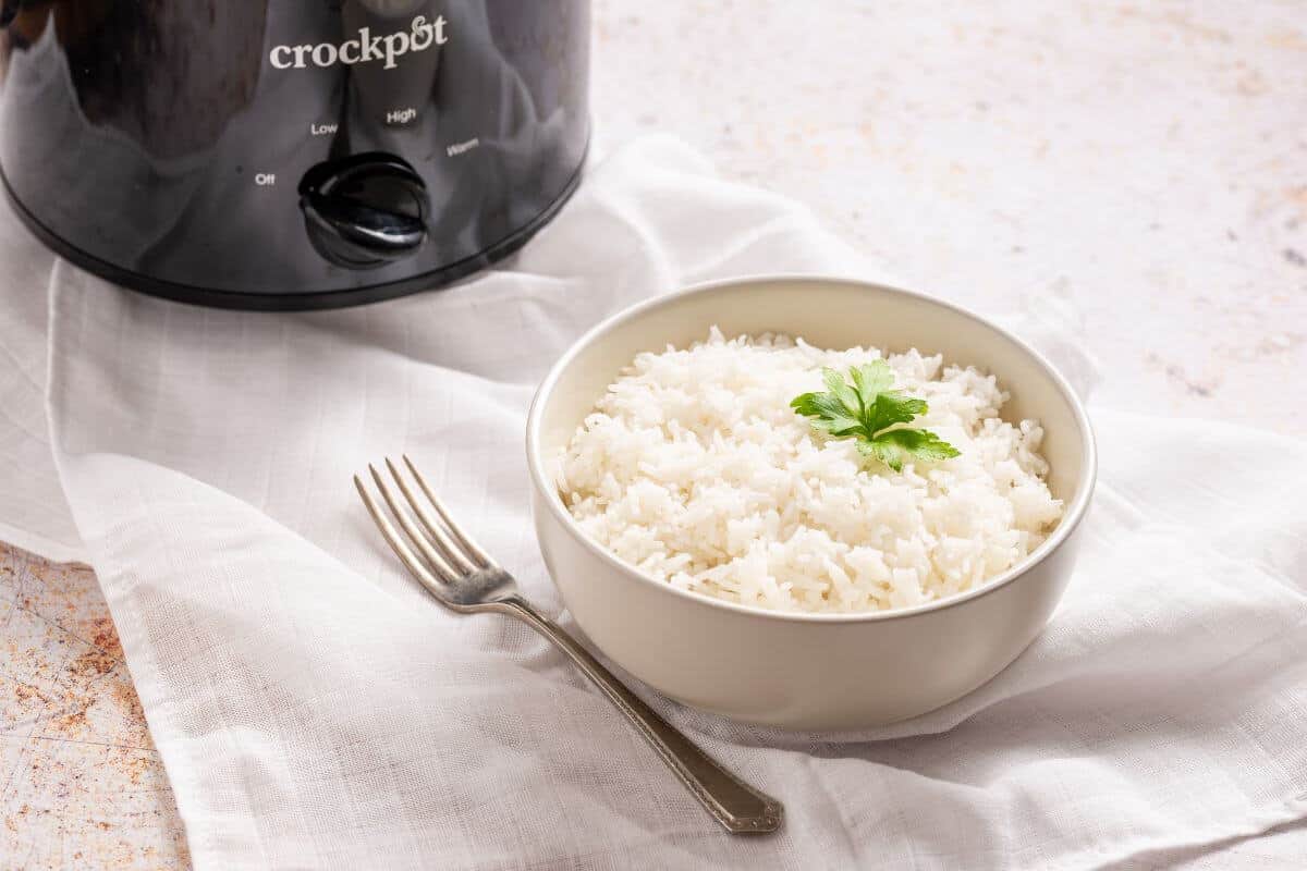 A bowl of rice with a fork and spoon in front of a slow cooker.