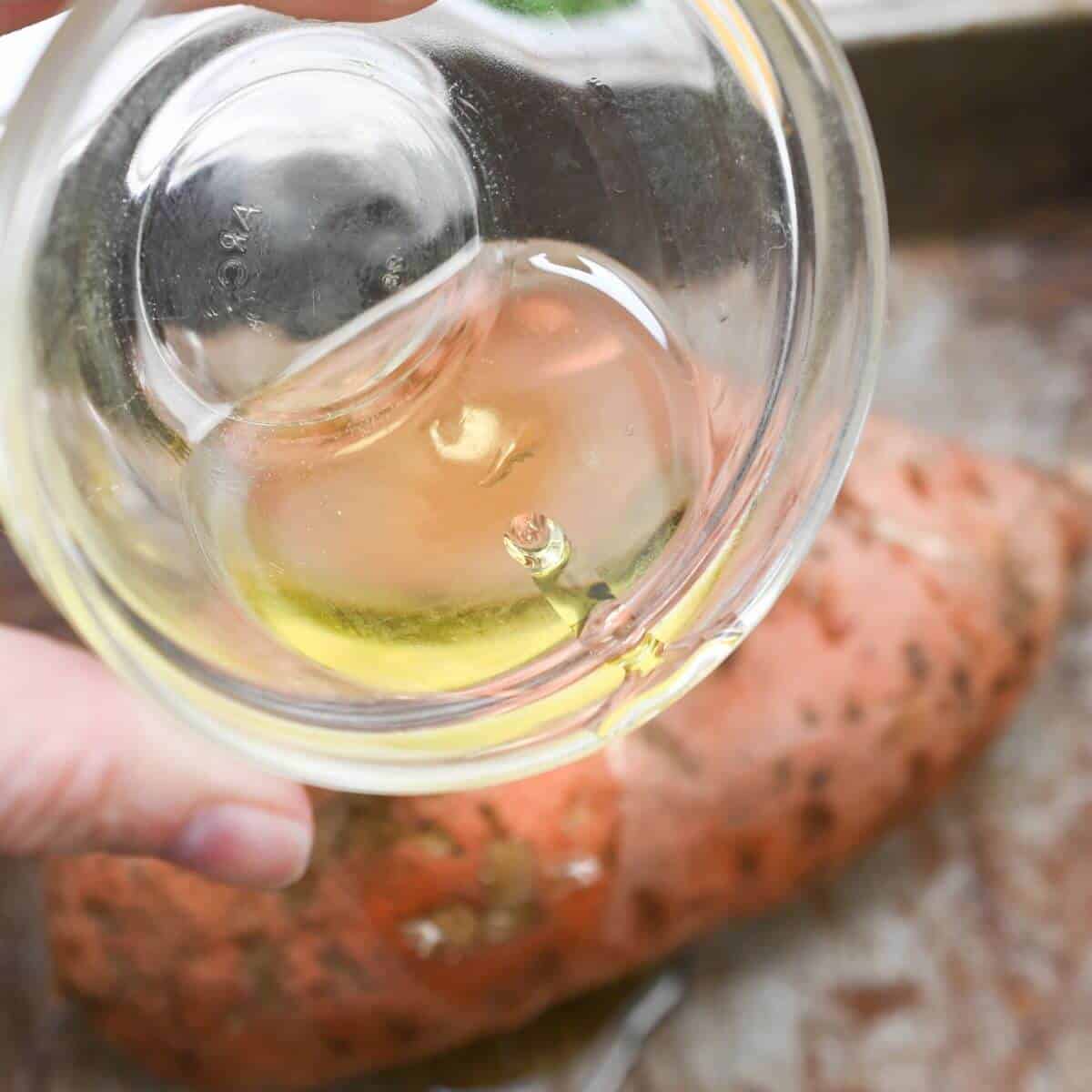 A person pouring oil into a glass next to a sweet potato.