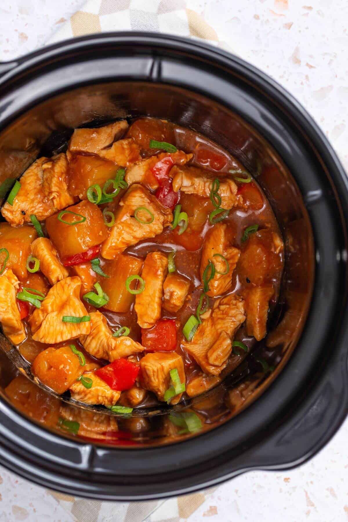 A crock pot filled with chicken, pineapple, and pepper in a sauce.